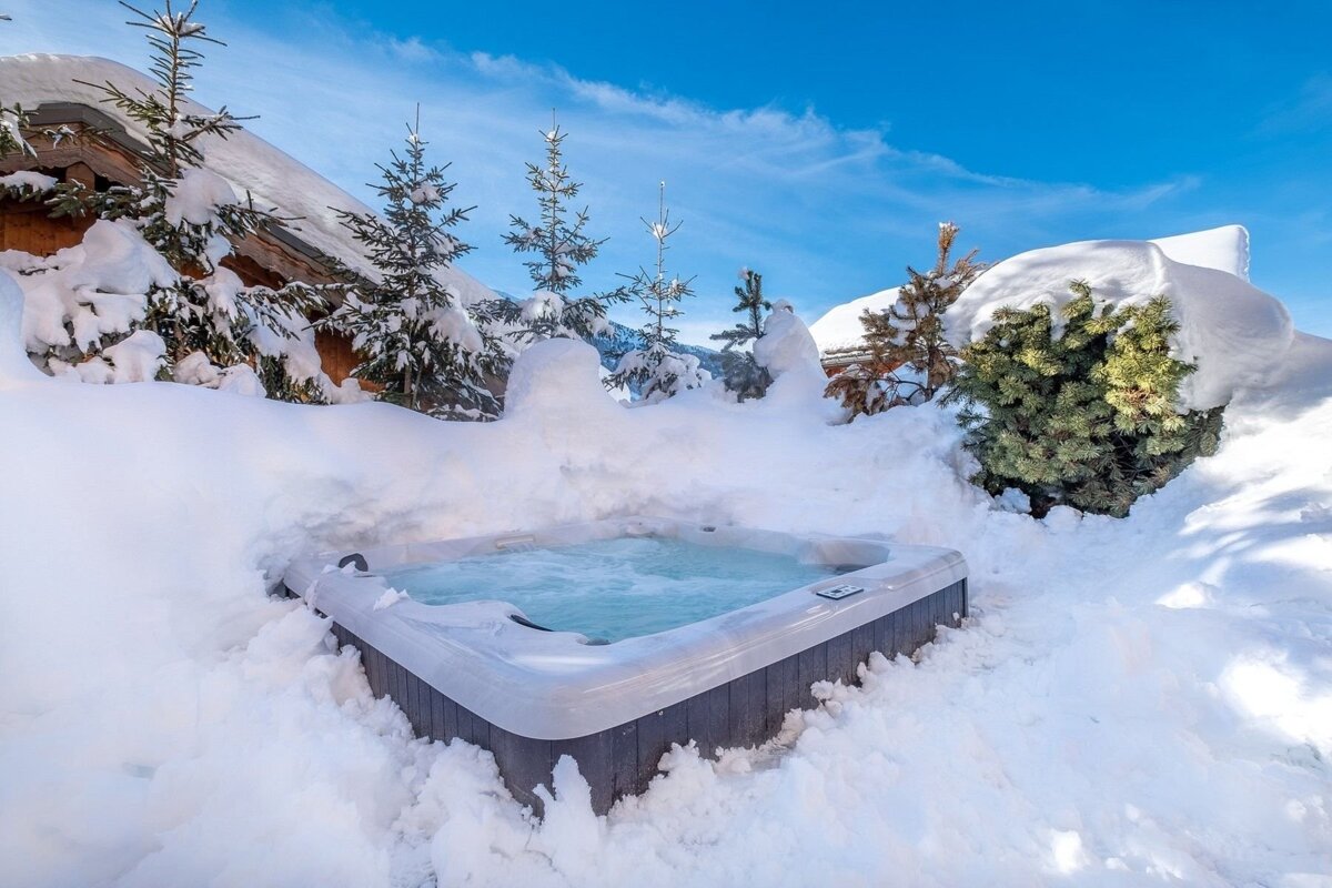 A steaming hot tub nestled in deep snow with snow-laden evergreen trees, a distant building, and a bright blue winter sky.