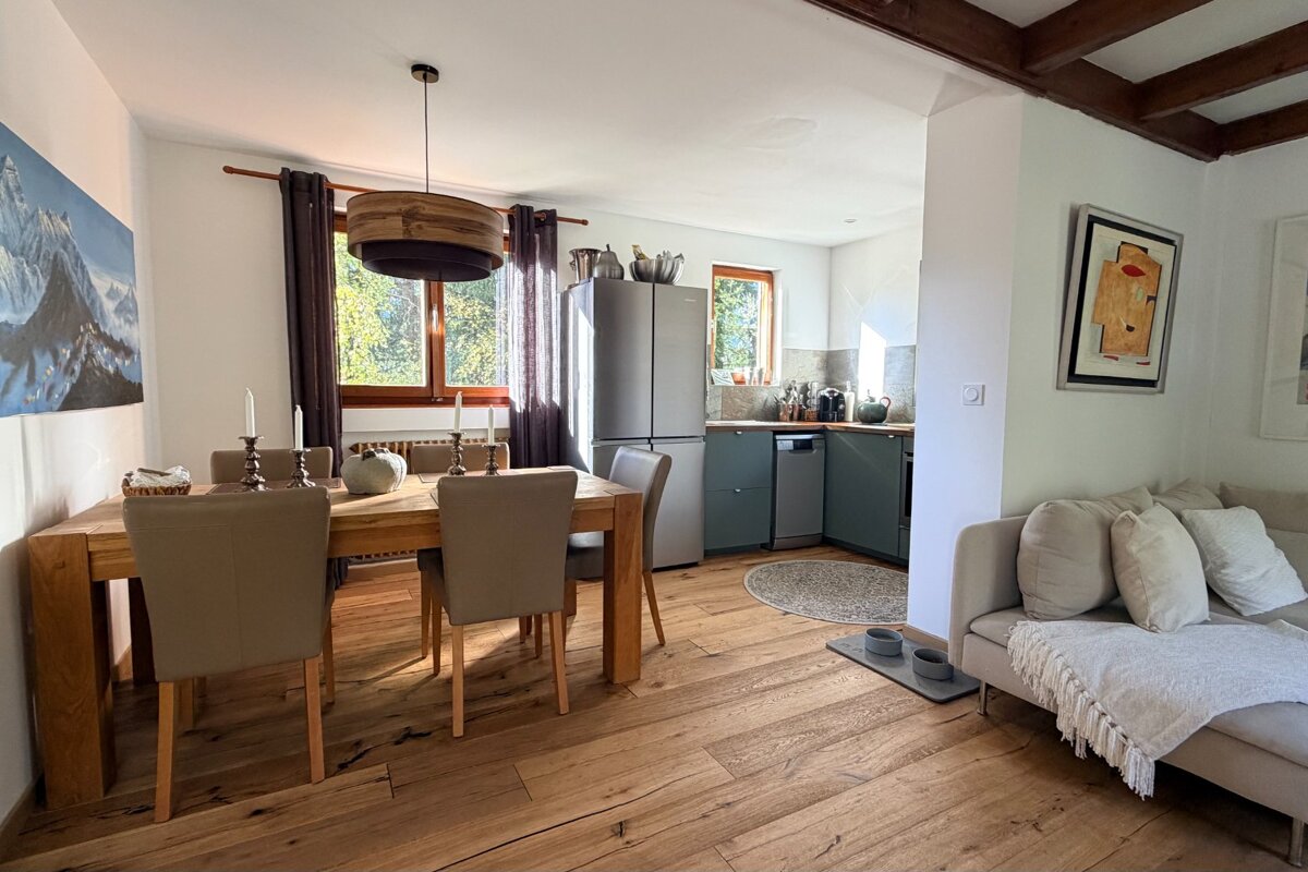 A bright, open-concept dining room, kitchen, and living area with warm wood floors, a large wooden dining table, green kitchen cabinets, and a cozy sofa.