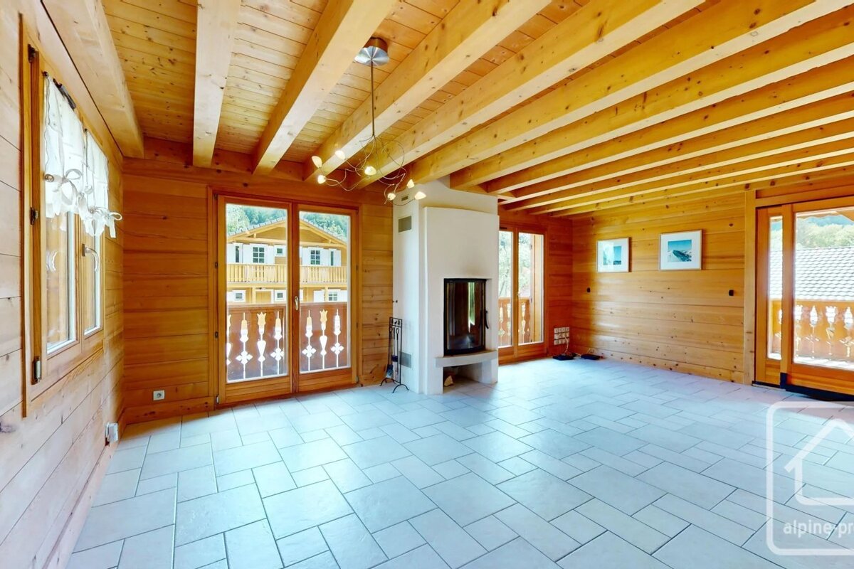 Empty wooden cabin interior with tiled floors, exposed ceiling beams, a white fireplace, and glass doors opening to balconies.