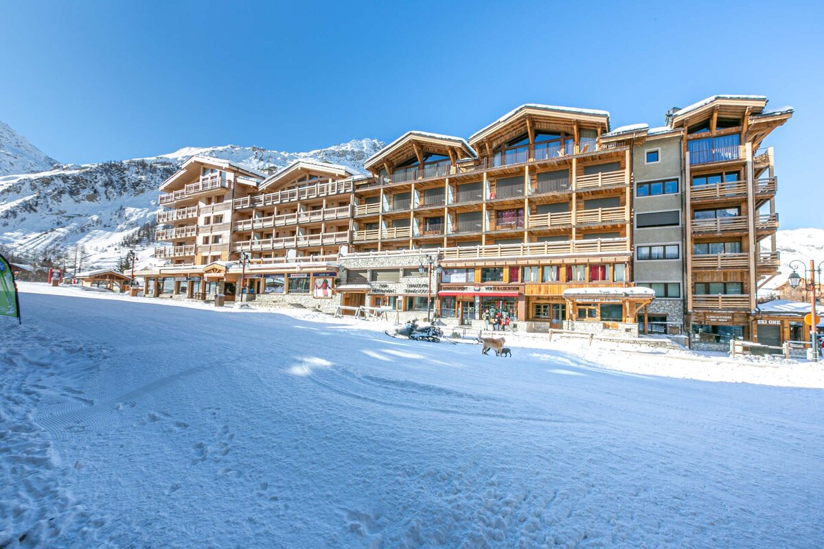 Multi-story, chalet-style buildings line a snowy ski slope at a mountain resort under a clear blue sky.