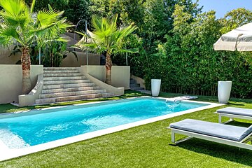 Sunny outdoor pool with artificial turf, palm trees, bamboo, stone stairs, and lounge chairs, creating a relaxing tropical setting.