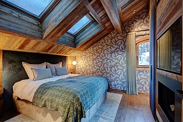 A cozy attic bedroom with a double bed, wooden ceiling beams, skylights, patterned wallpaper, and a window showing a snowy view.