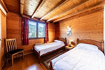 A cozy, wood-paneled bedroom features two twin beds with white bedding, red curtains, and a window offering a scenic mountain view.