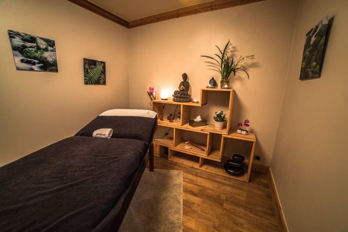 A tranquil spa room with a dark massage bed, wooden shelf displaying Buddha statues and plants, and warm ambient lighting. Nature art hangs on the walls.