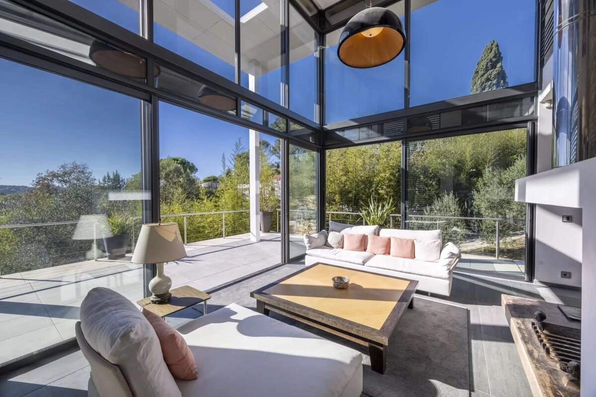 A sleek, modern living room bathed in natural light, featuring expansive glass walls that showcase a stunning, vibrant green natural landscape.