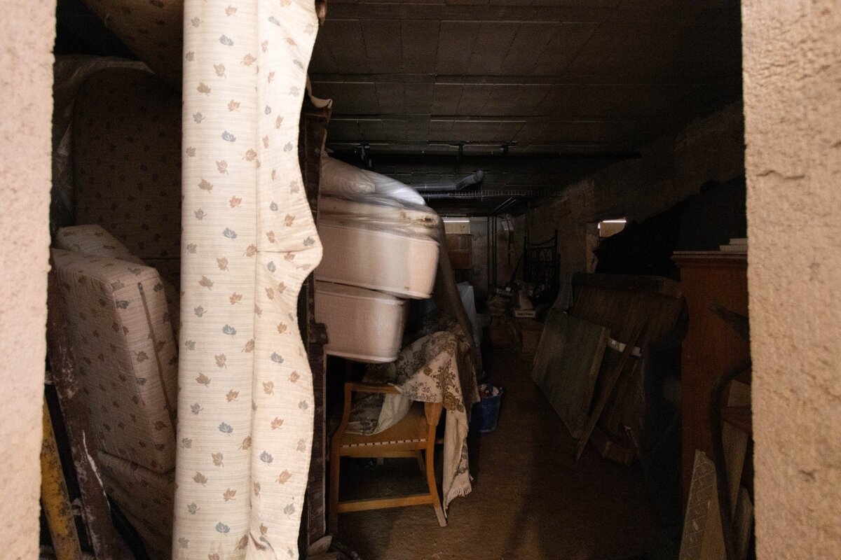 A dark, narrow storage space packed with old mattresses, furniture, and boxes, viewed through a gap in patterned fabric.