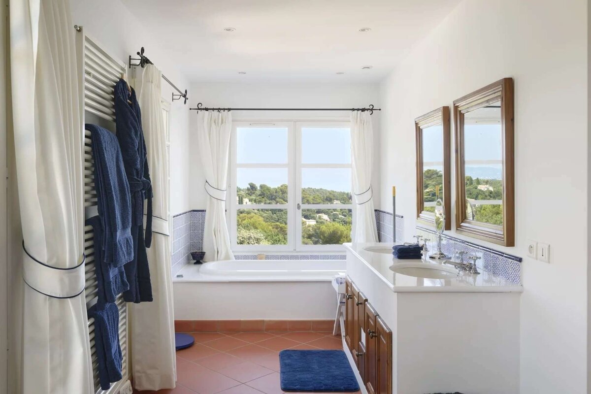 A bright bathroom with a tub by a scenic window, a double vanity, terracotta floor, and blue accents on towels, bathmat, and curtain trim.