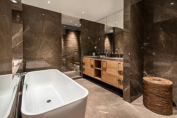 A luxurious modern bathroom featuring dark marble walls, a freestanding white tub, wooden double vanity, walk-in shower, and woven hamper.