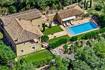 Aerial view of a large, rustic villa with terracotta roofs, a blue pool, and extensive green gardens, surrounded by lush trees and a natural, hilly landscape.