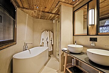 Cozy, modern bathroom with a wooden ceiling, freestanding tub, double vessel sinks, and hanging robes, evoking a rustic-chic chalet style.