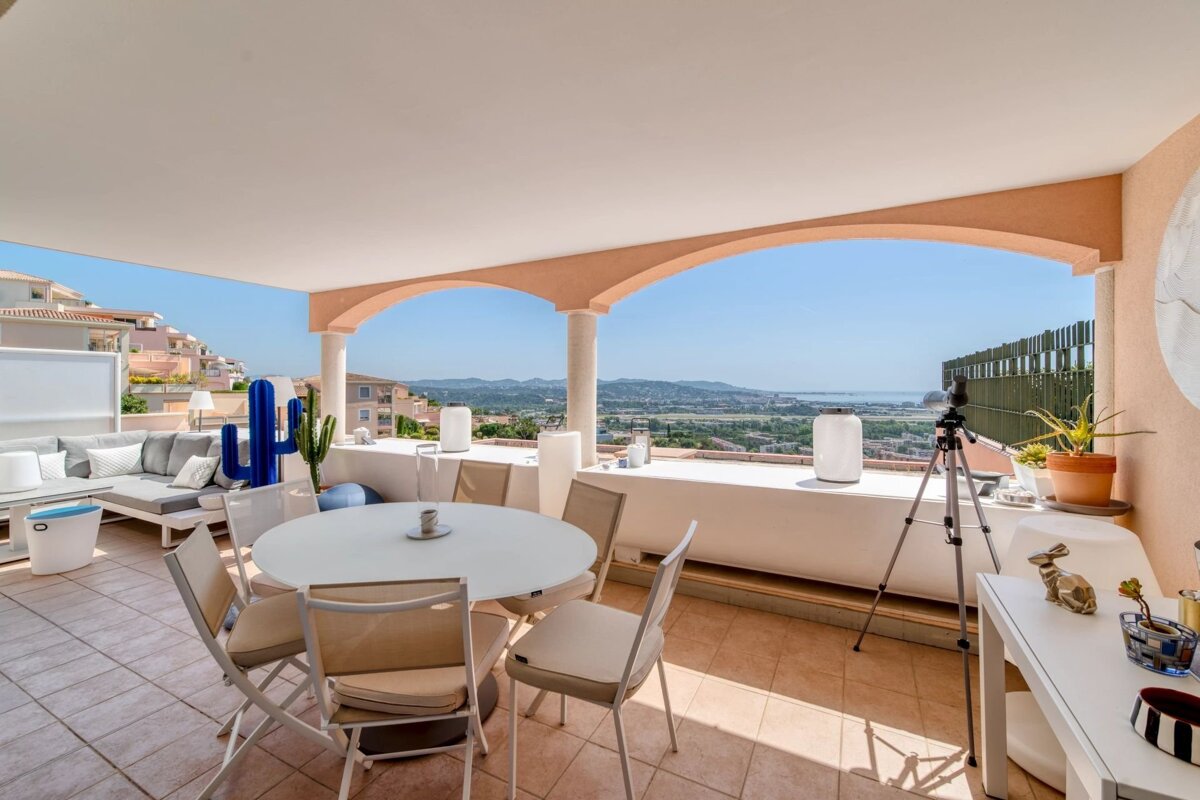 Modern covered terrace with dining and lounge seating, a blue cactus, and arched views of a distant landscape and sea.