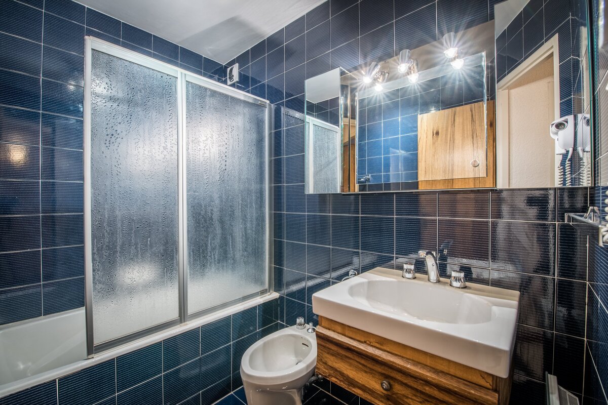 A blue tiled bathroom featuring a tub/shower with frosted glass, a white sink on a wooden vanity, a bidet, and a mirror with bright overhead lights.