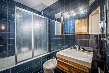 A blue-tiled bathroom with a steamy shower over a tub, a white sink on a wooden vanity, a bidet, and a brightly lit mirror.
