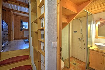 A multi-level wooden interior featuring stairs, a built-in bookshelf, and a compact bathroom with a shower and toilet nestled beside it.