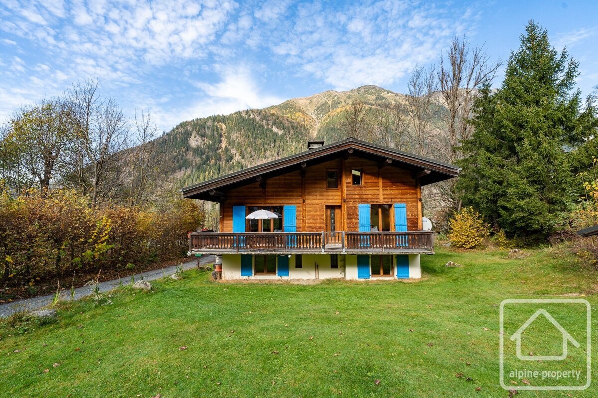A charming wooden chalet with blue shutters stands on a green lawn, backed by mountains and autumn trees under a clear blue sky.