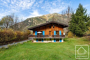 A charming wooden chalet with blue shutters stands on a green lawn, backed by mountains and autumn trees under a clear blue sky.