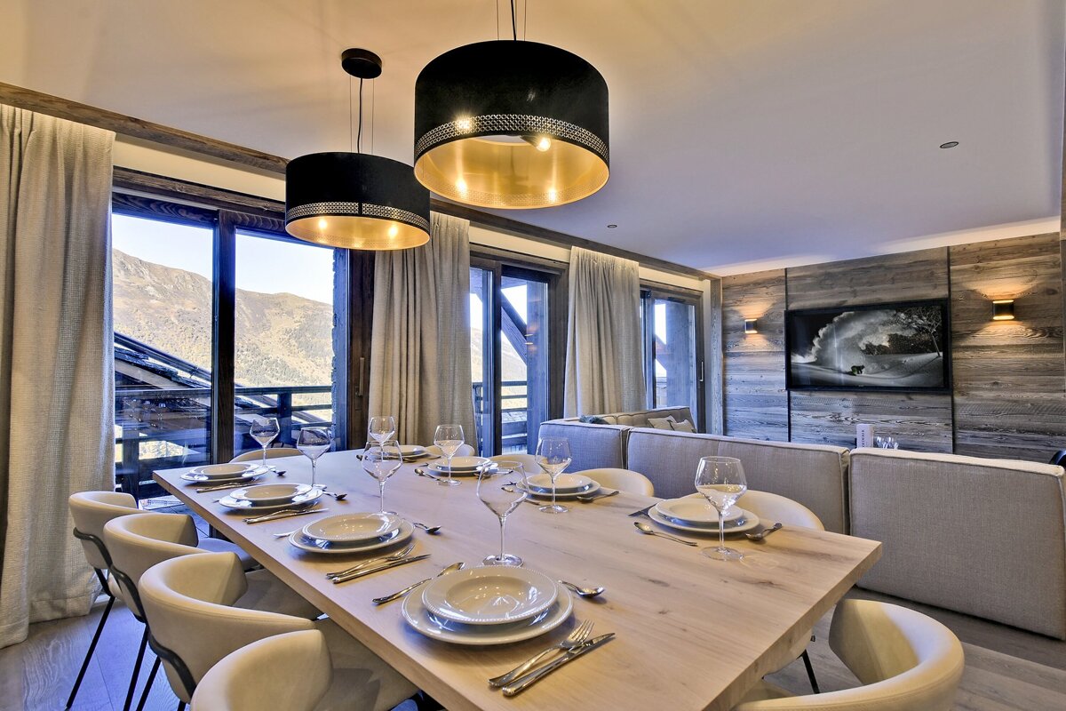 A stylish dining room with a large wooden table set for dinner, featuring modern black and gold lights, large windows overlooking a mountain view, and a cozy living area.