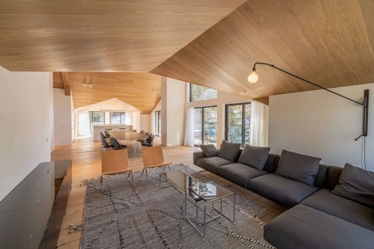 A spacious, modern open-plan living area featuring warm wooden vaulted ceilings and floors, a large grey sectional sofa, and floor-to-ceiling windows.
