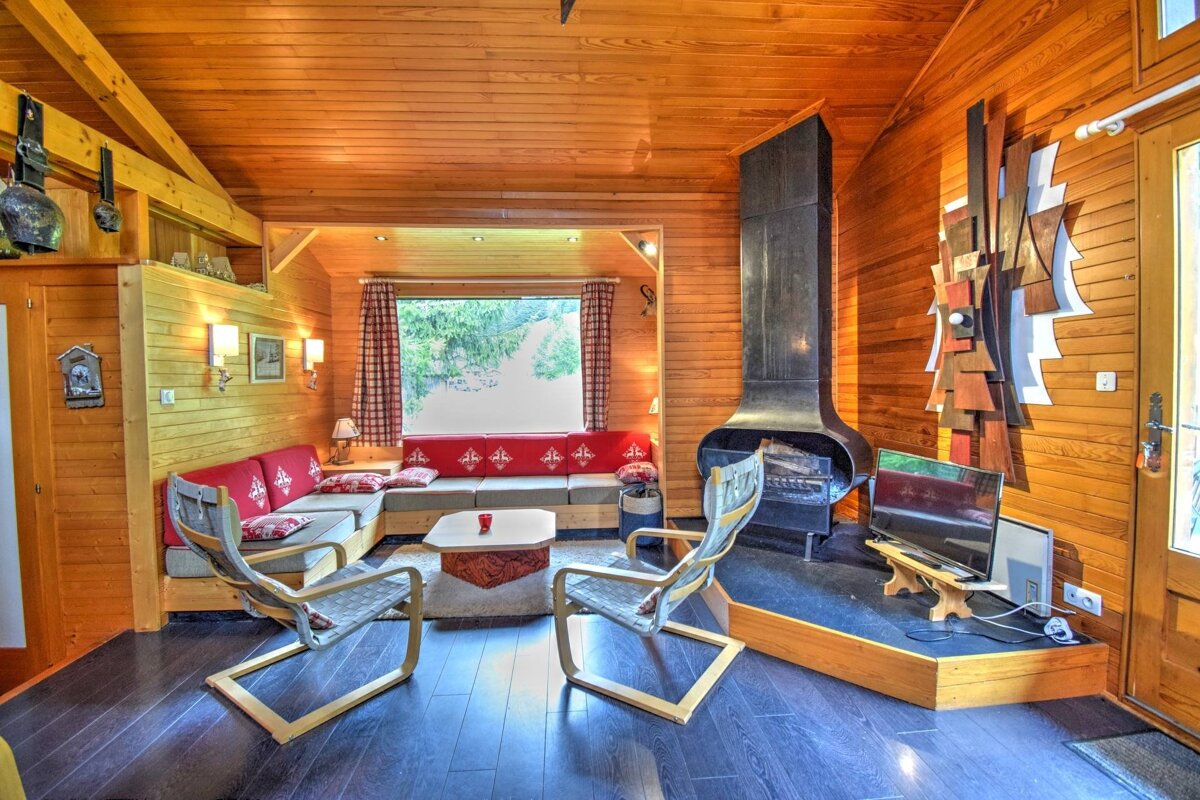Cozy, wood-paneled living room with a large fireplace, red sectional sofa, modern chairs, and a window overlooking nature.