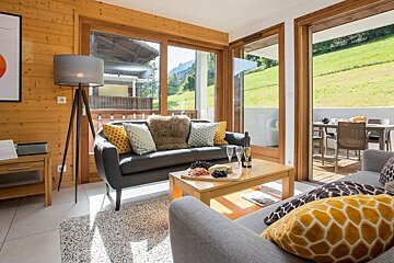 Cozy chalet living room featuring grey sofas, a wooden coffee table, and large windows overlooking a green mountain hillside.