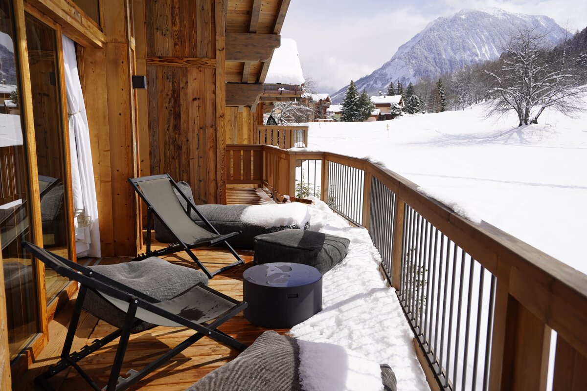 A wooden chalet balcony with lounge chairs looks out over a snow-covered mountain valley.