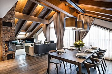 A stylish open-plan living and dining room with a vaulted wooden beam ceiling, stone fireplace, modern furnishings, and large bright windows.