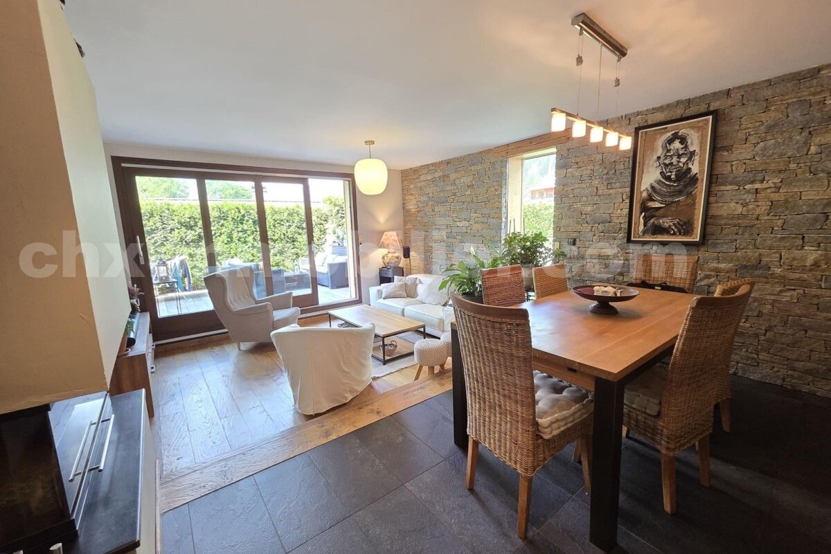 Spacious living/dining room with a rustic stone accent wall, modern light-colored furniture, a wooden dining table, and large glass doors to an outdoor patio.