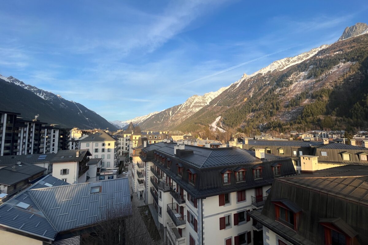 Rooftop view of an alpine town with traditional buildings, flanked by majestic snow-capped mountains under a clear, sunny blue sky.