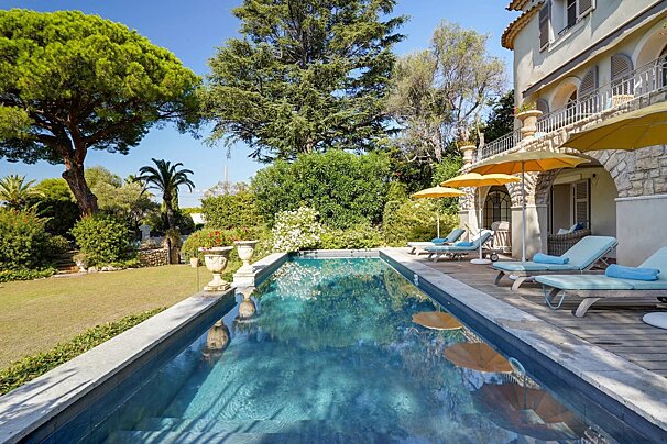 A sunny backyard pool with blue loungers, yellow umbrellas, and a stone villa surrounded by lush green trees.
