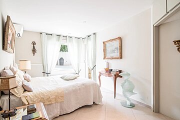 Elegant, bright white bedroom with a large bed, sheer curtains, and a window looking out onto a pool. Classic furniture and artwork.