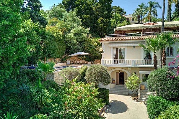 A luxurious cream villa with balconies and a tiled roof is surrounded by abundant green trees and foliage, featuring a sun-drenched pool area.