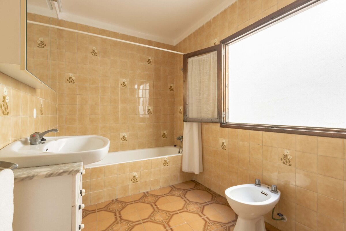 A tan-tiled bathroom with floral patterns, featuring a bathtub, sink, bidet, and a large window. The floor has octagonal tiles.