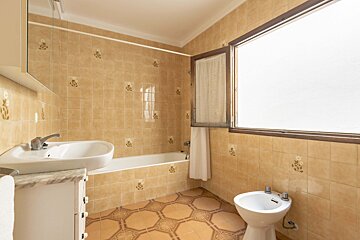 A tan-tiled bathroom with floral patterns, featuring a bathtub, sink, bidet, and a large window. The floor has octagonal tiles.