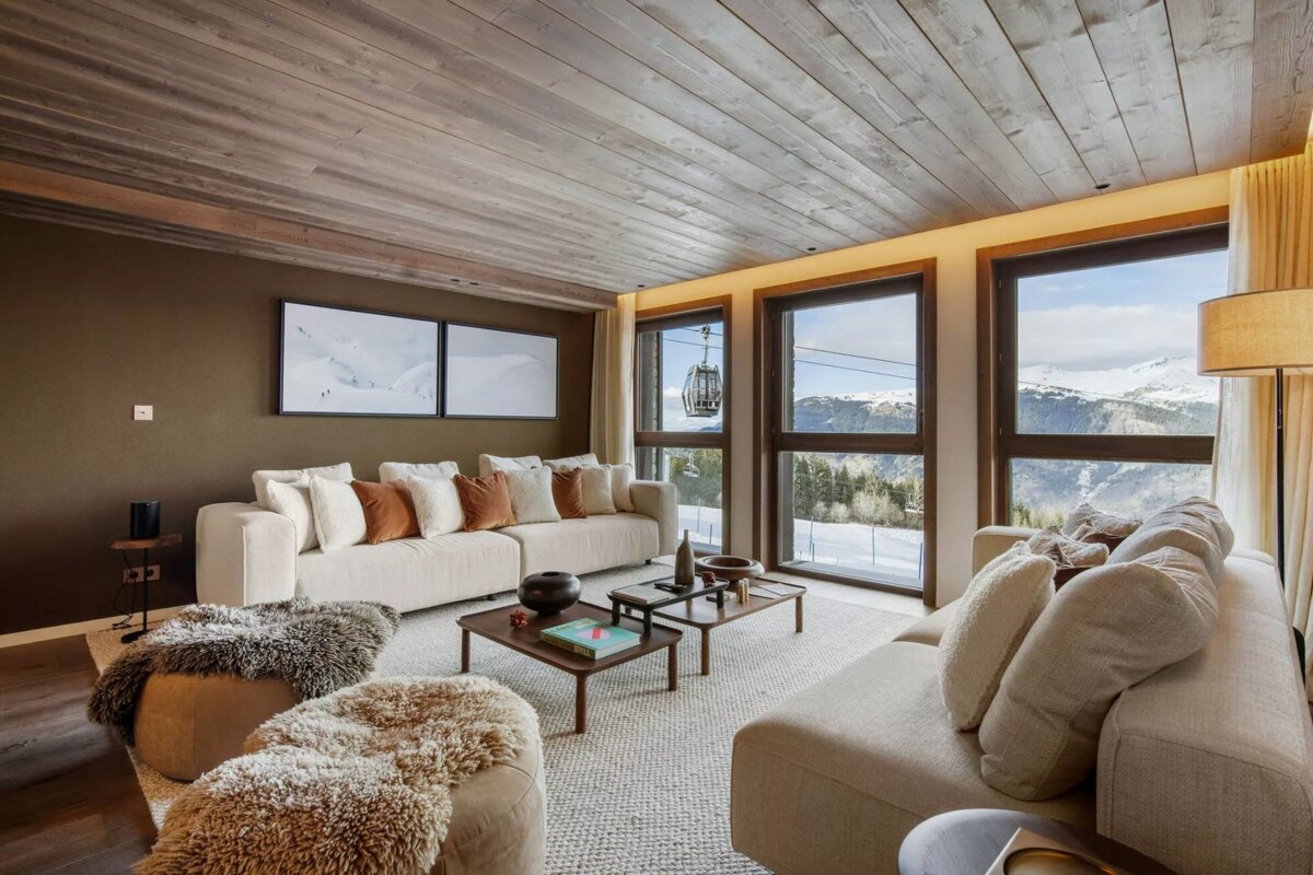 Chic ski chalet living room with plush seating, wood ceiling, and floor-to-ceiling windows revealing a snowy mountain vista and ski lift.