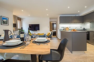 Modern open-plan apartment featuring a set wooden dining table, a cozy living room, and a sleek grey kitchen.