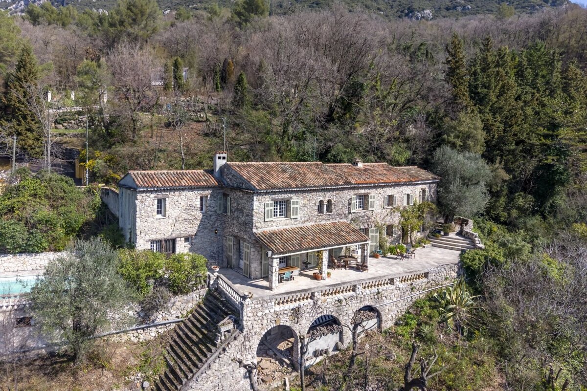 Large stone house with terracotta roof tiles on a terraced hillside amidst dense woods. Features several patios and a distant swimming pool.