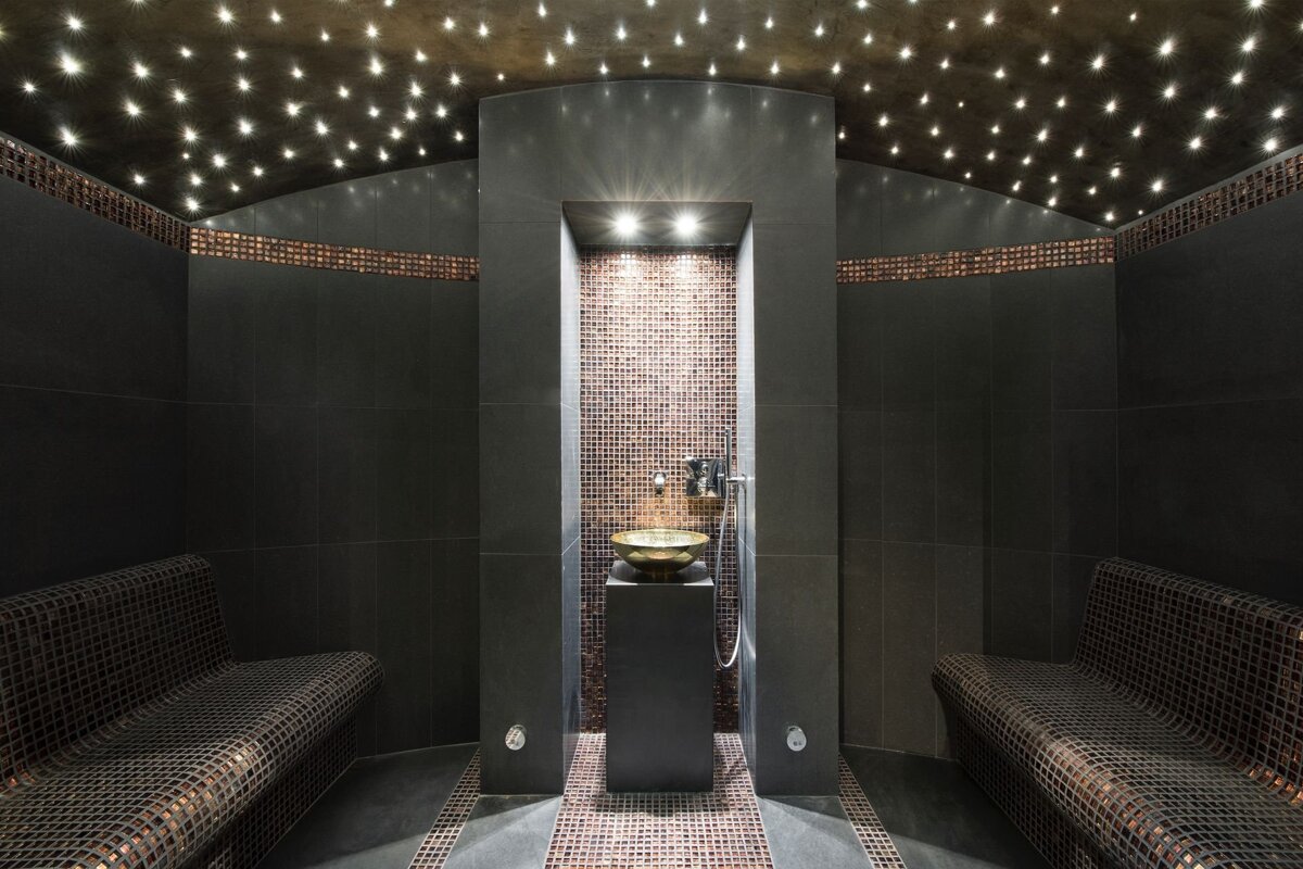 A luxurious dark-tiled spa or hammam with a starry ceiling, mosaic-covered benches, and a central golden bowl for ritual washing.