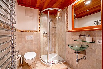 A compact bathroom with a corner glass shower, glass vessel sink, wall-mounted toilet, chrome towel rack, and a warm wooden ceiling.
