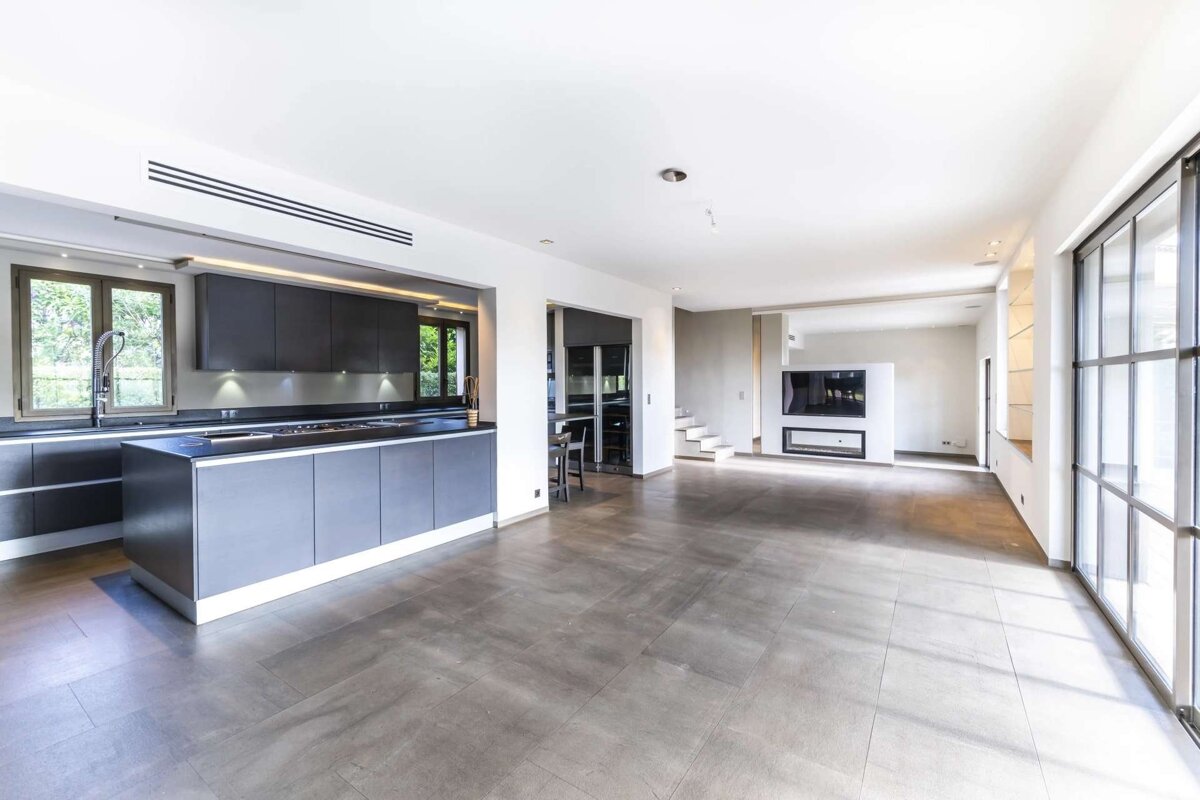 Modern open-plan living space with dark grey kitchen, grey tiled floor, large windows, and a living area with a TV and fireplace.