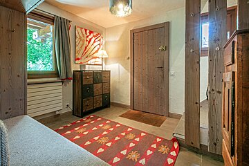 A cozy, rustic room with a wooden door, a vibrant red patterned rug, a multi-drawer cabinet, and a window looking out to green trees.