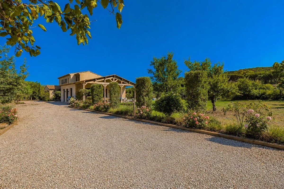 A scenic view of a country house with a wooden pergola, a wide gravel driveway, lush gardens with pink roses, under a clear blue sky.