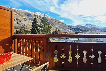 A wooden balcony with ornate railings overlooks snow-dusted mountains, green hills, and evergreen trees under a bright blue sky.