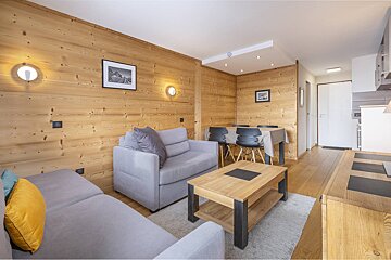 Wood-paneled living/dining room with gray sectional sofa, wooden coffee table, and dining table with black chairs. An open-plan kitchen is to the right.