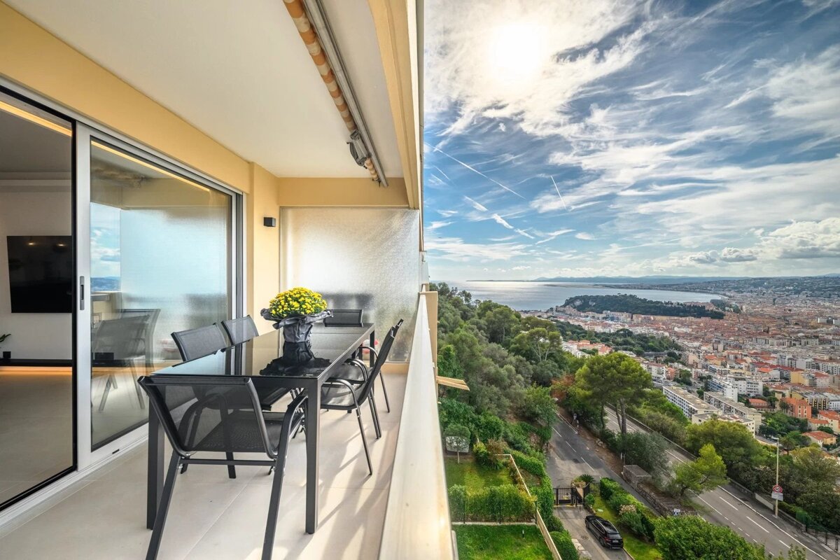 Balcony dining table overlooking a coastal city and sea under a bright, cloudy sky.