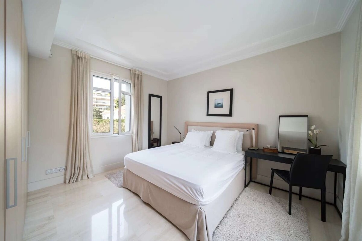 A bright, neutral-toned modern bedroom with a white bed, large window, sleek desk, and wardrobe. Clean design and polished light floors.