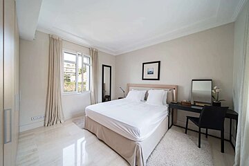 A minimalist modern bedroom features a bed with white linens, a large window, a full-length mirror, and a dark desk. Light walls and marble floor.