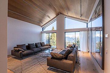 A modern living room with large panoramic windows overlooking snowy mountains. It features sectional sofas, a geometric rug, and a striking wooden vaulted ceiling.