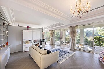 A bright, elegant living room with a white sofa, blue rug, and ornate chandelier. Large glass doors open to a spacious balcony with a lush, green view.