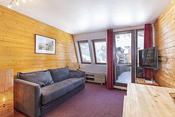 A cozy, wood-paneled ski apartment living room with a grey sofa, purple carpet, and windows overlooking a snowy mountain village.