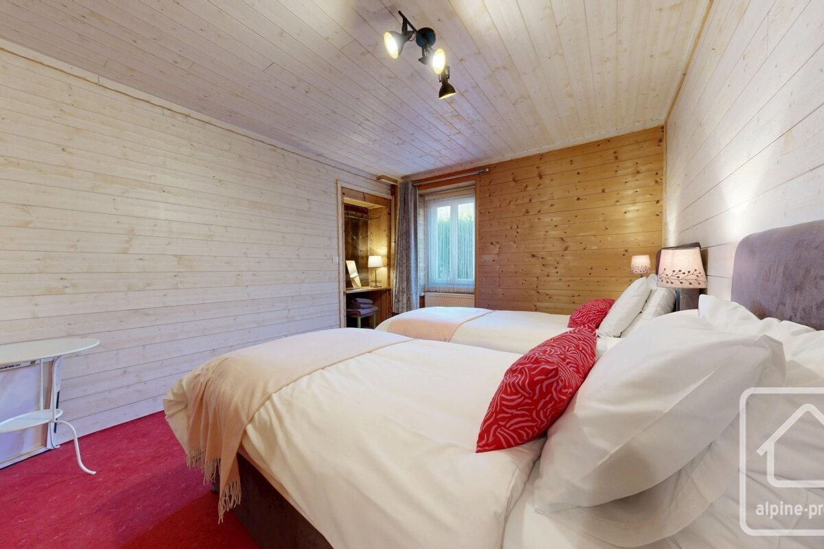Cozy bedroom with two single beds, light and dark wood-paneled walls, red carpet, and a window. Features bedside lamps and ceiling lights.